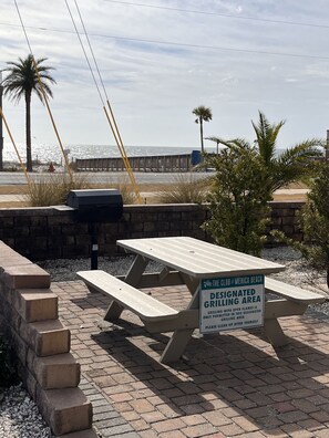 Zona de barbacoa designada con mesas de picnic y una parrilla, cerca de una playa con palmeras y un muelle al fondo.