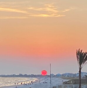 Escena de playa con atardecer, gente caminando por la orilla y edificios al fondo.