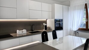 A modern kitchen with white cabinetry, a black countertop, and a dining area with a white table and black chairs. - Spacious 95 sqm newly renovated vacation home with breathtaking views (Menaggio)