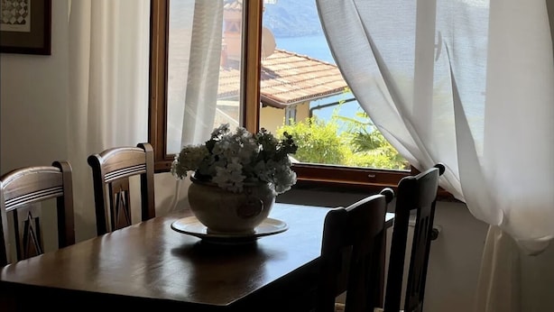 A dining table with a bowl of flowers, wooden chairs, and a window with sheer curtains. - Spacious 95 sqm newly renovated vacation home with breathtaking views (Menaggio)