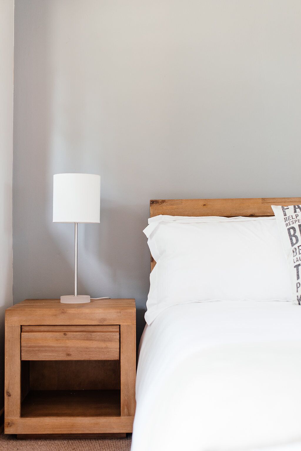 A bed with white bedding and a wooden headboard, a white bedside lamp on a wooden nightstand, and a plain wall.