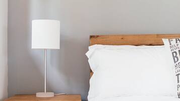 A bed with white bedding and a wooden headboard, a white bedside lamp on a wooden nightstand, and a plain wall.
