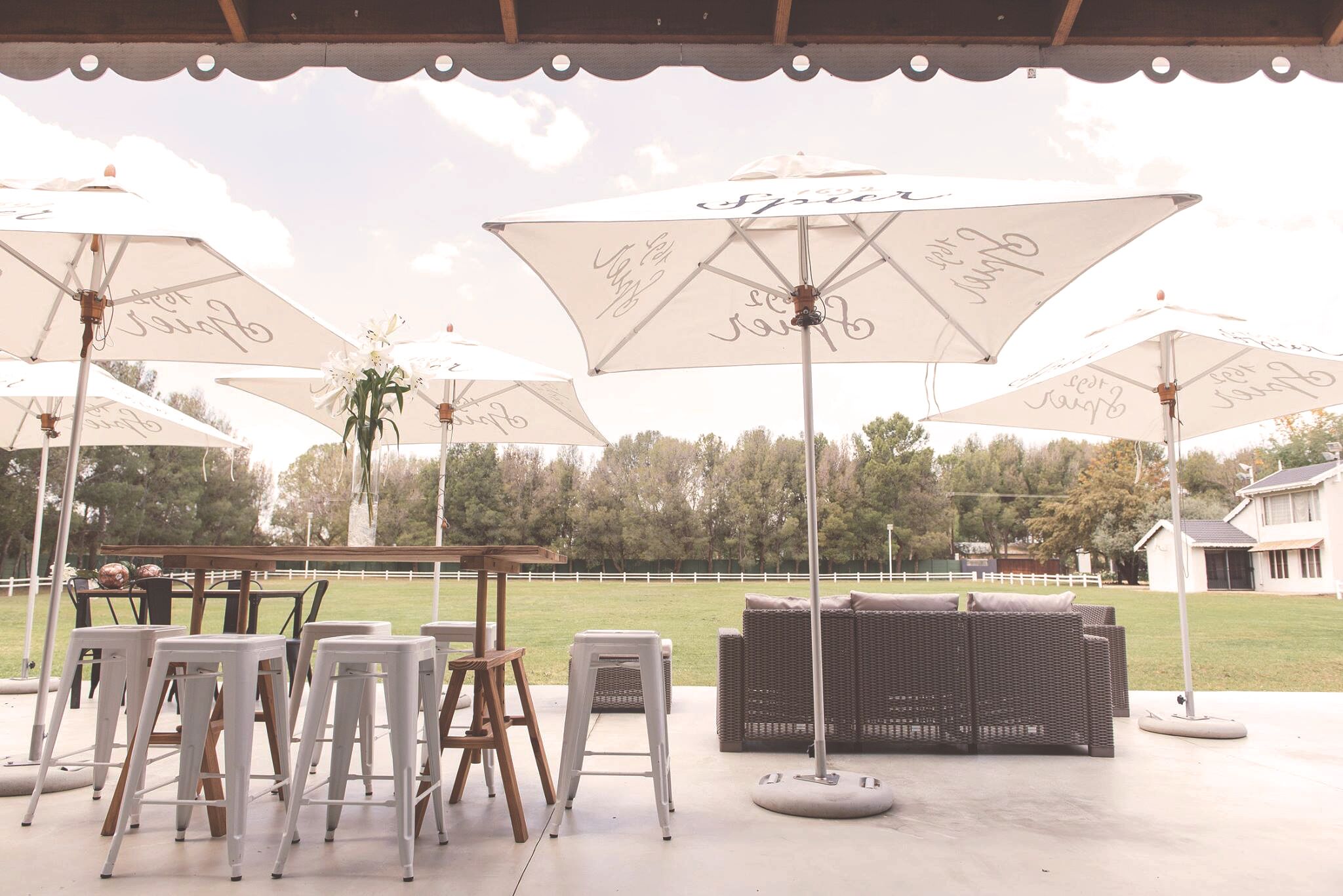 An outdoor patio area with white umbrellas, wicker furniture, and a grassy field in the background.
