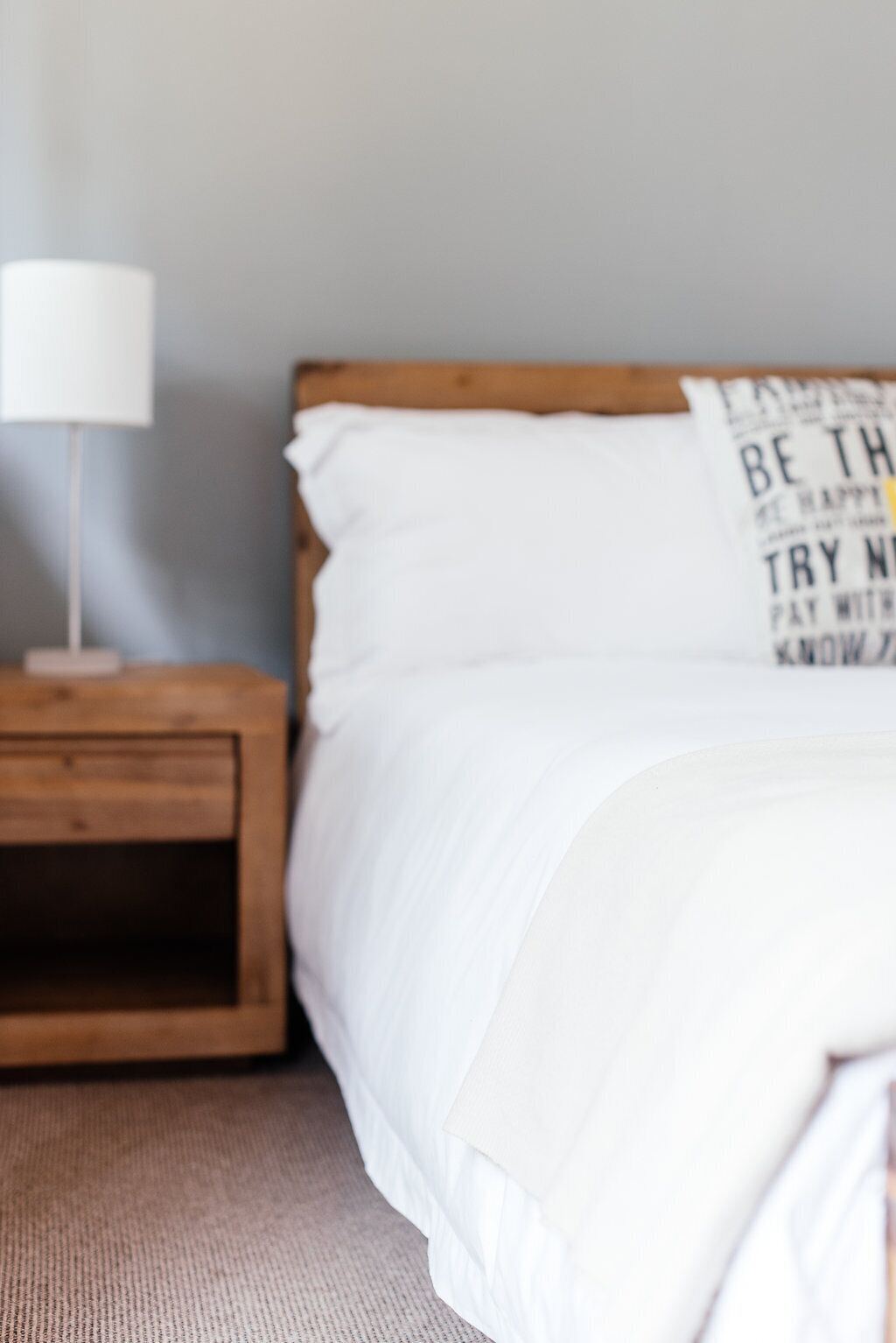 Een slaapkamer met een houten nachtkastje, een witte lamp en een bed met witte lakens.