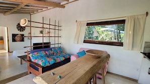 A cozy room with a wooden bench, a sofa with colorful cushions, a rustic wall hanging, and a window with curtains.
