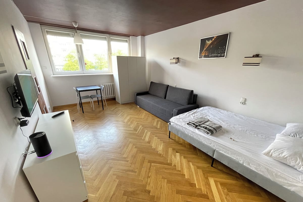 A room with a bed, a grey sofa, a small table with chairs, a desk with a computer, and a window with blinds.