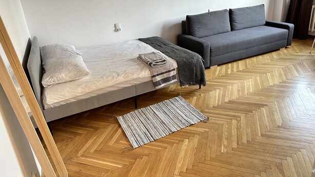 Un dormitorio moderno con cama, un sofá gris y suelo de madera con diseño de espiga.