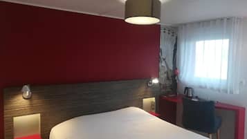 Una habitación de hotel con cama, escritorio, silla, lámpara y ventana con cortinas.