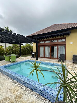 A swimming pool with a tiled edge, surrounded by a pergola, outdoor furniture, and a building with a tiled roof and glass doors. - Tranquil Bungalow w Pool at Green Village Ca (Punta Cana)