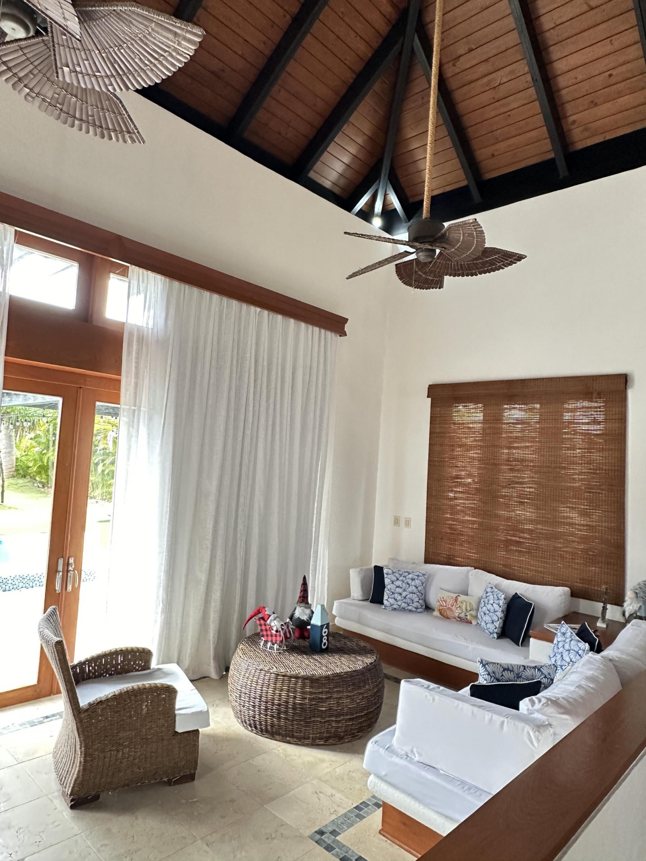 A spacious living area with a high ceiling, a ceiling fan, a sofa set, a wicker chair, and a large window with curtains.