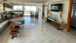 A spacious living area with a sofa, coffee table, and TV. There is a dining area with chairs and a table. The room has a tiled floor and a large window. - Dar Riahi Ras Jbal (Ras Jebel)
