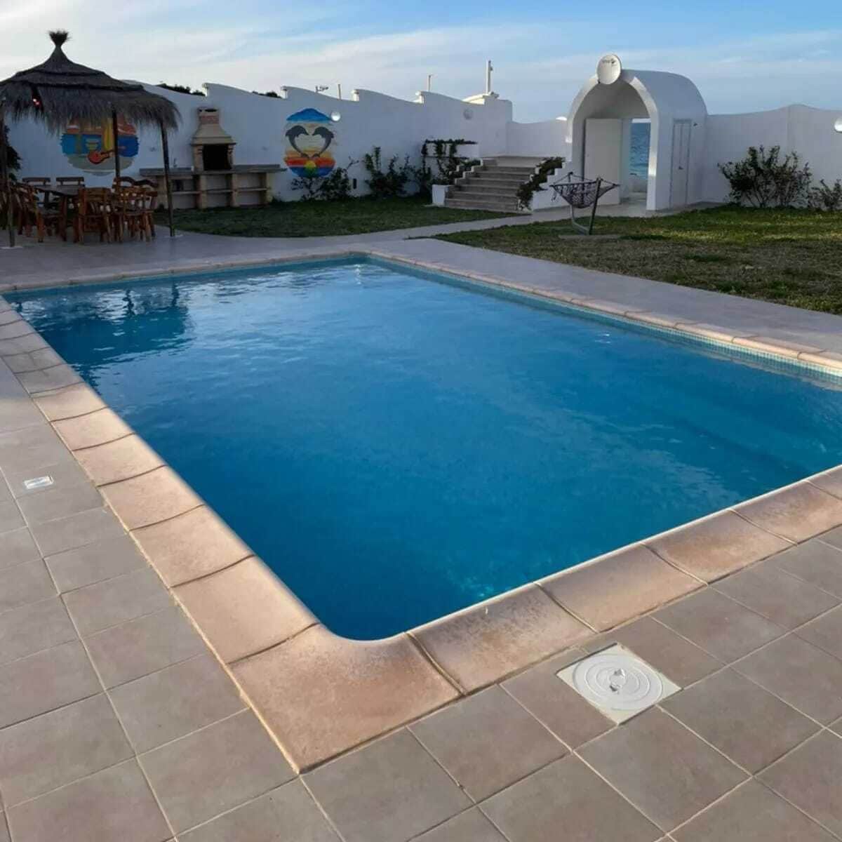 A rectangular swimming pool with a tiled edge, surrounded by a grassy area and a white building with a mural.
