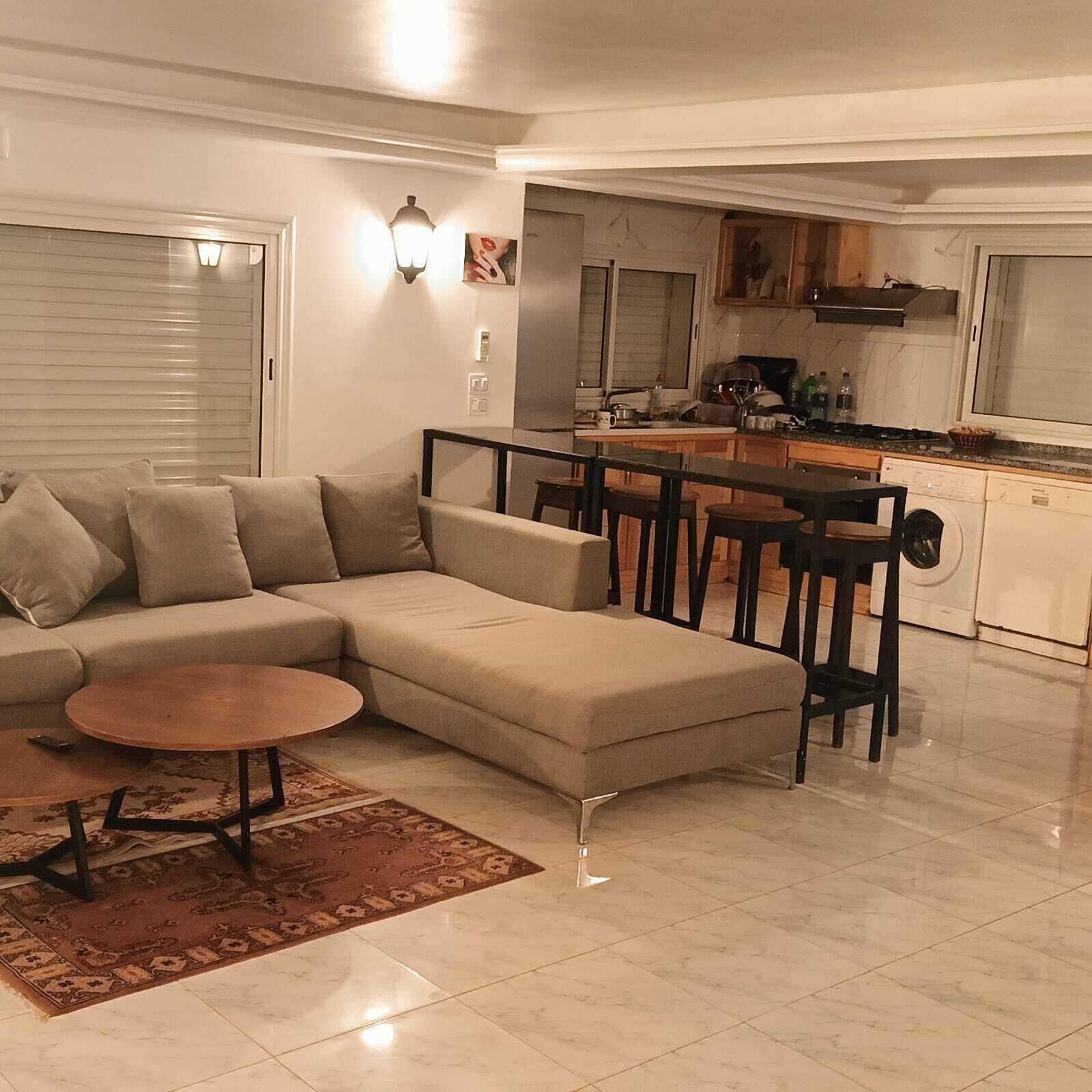 Una sala moderna con un sofá seccional color beige, una mesa de centro redonda y un área de cocina con una mesa de comedor y taburetes de bar.