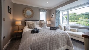 A bedroom with a large bed, a bedside table with a lamp, a window with a view of greenery, and a circular mirror on the wall.