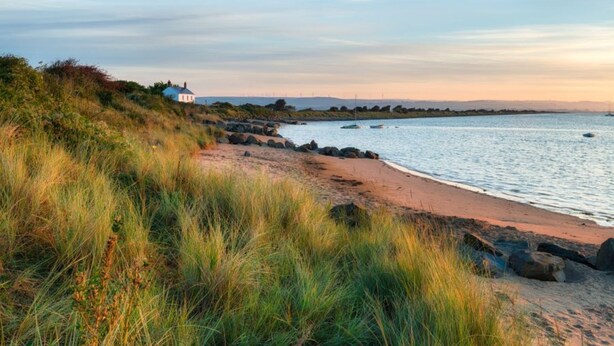 A coastal landscape with a sandy beach, grassy dunes, and a small building near the water.