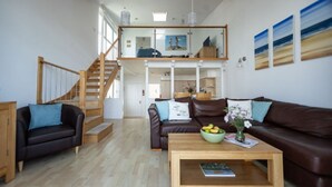 A spacious living area with a wooden staircase, a brown leather sofa, a wooden coffee table with a bowl of fruit, and a view of the upper floor through a glass door. - 2 Bed Beach House in a fantastic sea front setting (Westward Ho!)