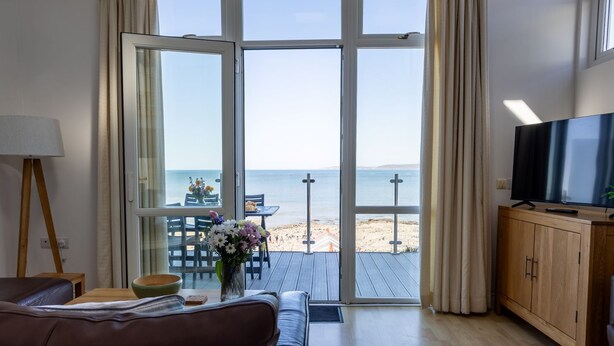 A room with a large sliding glass door leading to a beach view, a wooden cabinet with a TV, a brown leather sofa, and a small table with a vase of flowers. - 2 Bed Beach House in a fantastic sea front setting (Westward Ho!)