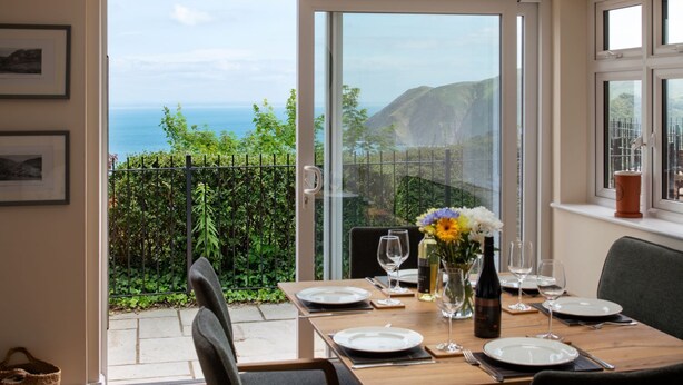 Un comedor con una mesa dispuesta para dos, con vistas panorámicas al mar y las colinas a través de grandes puertas de vidrio.