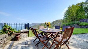 Un patio exterior con una mesa redonda y sillas de madera, un jarrón con flores y vistas al mar y las colinas.