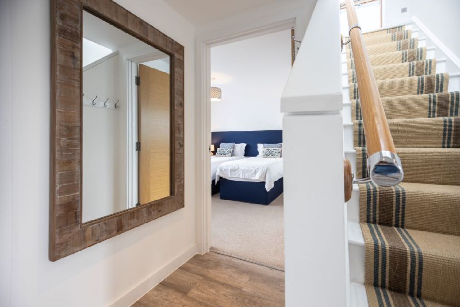 Ein Flur, der zu einem Schlafzimmer führt, mit einem großen Spiegel, Holzboden und einer Treppe mit Streifenmuster.