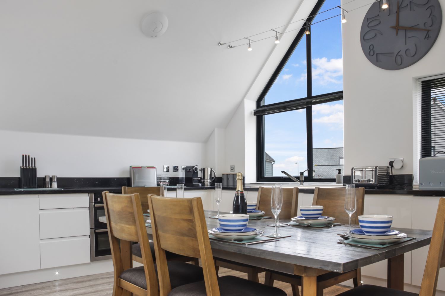 A modern kitchen with a dining area, featuring a large window, a clock, and a dining table set for four.