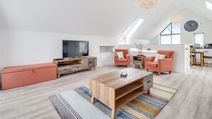 Un salon spacieux avec un plafond haut, un grand téléviseur, une table basse en bois et deux fauteuils roses.