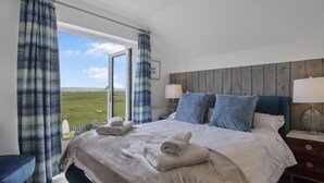 A bedroom with a large bed, two bedside lamps, a view of the outdoors, and a wooden headboard. - Luxury 4 bed beachside home with coastal views (Westward Ho!)