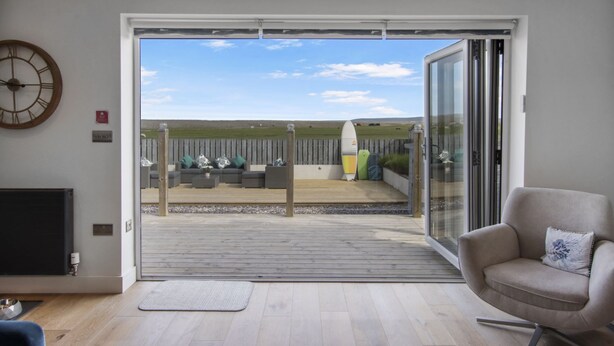 A modern living room with a large sliding glass door leading to an outdoor deck with seating and surfboards. - Luxury 4 bed beachside home with coastal views (Westward Ho!)