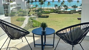 A balcony with two black chairs and a small table, overlooking a well-maintained lawn, palm trees, and a beach with a clear blue sky. - Modern Studio With Ocean View in Punta Palmera (Punta Cana)