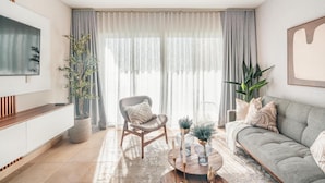 A modern living room with a grey sofa, a wooden coffee table, and a grey chair. There is a large window with sheer curtains, a flat-screen TV mounted on the wall, and a potted plant. - Modern Studio With Ocean View in Punta Palmera (Punta Cana)