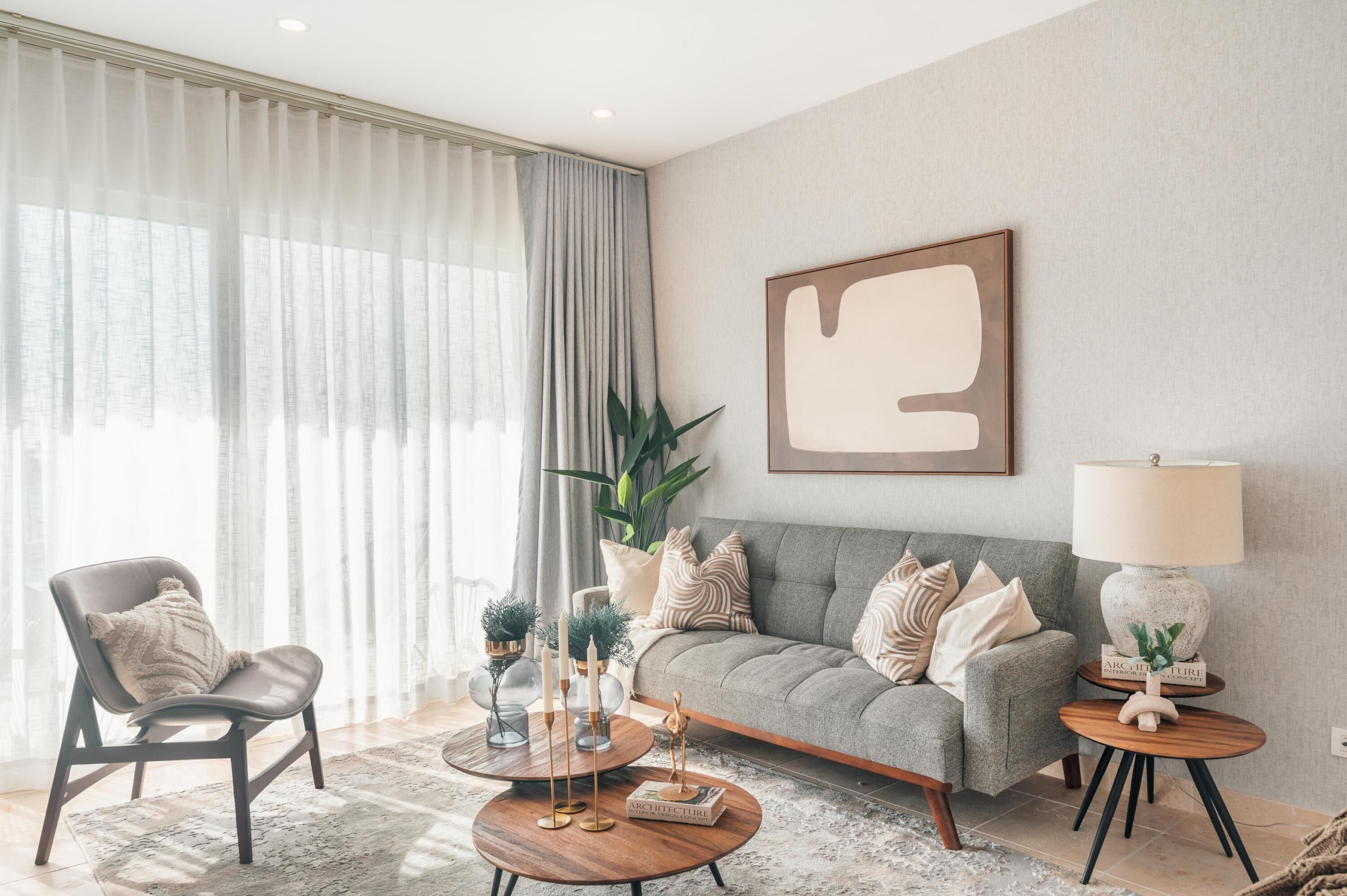 A modern living room with a grey sofa, a round coffee table, and a chair. There is a large window with sheer curtains, a potted plant, and a framed artwork on the wall.