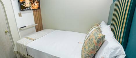 A single bed with a white mattress and colorful pillows, a wooden headboard, a mounted TV on the wall, and a door with a light switch.