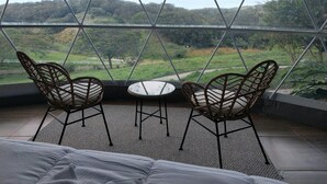 A room with a large window offering a view of a hilly landscape. There are two wicker chairs and a small round table in the room. - Vida Aventura Glamping Deluxe Dome (Bagaces)