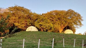 A grassy hill with two dome-shaped structures, surrounded by trees and a clear sky. - Vida Aventura Glamping Deluxe Dome (Bagaces)