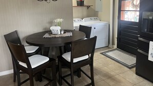 Un comedor con una mesa redonda, cuatro sillas, una araña, una lavadora y un refrigerador.