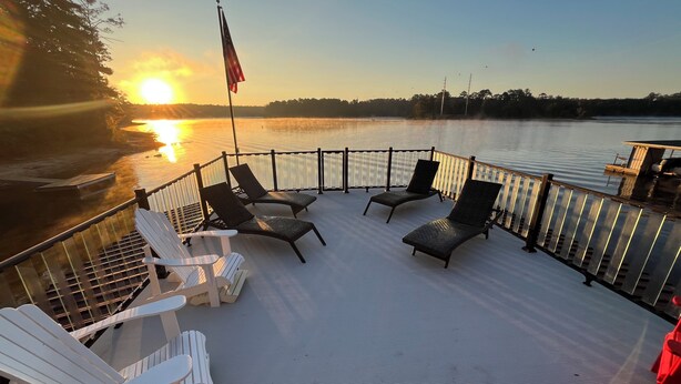 Una terraza con sillones reclinables y sillas Adirondack con vista a un cuerpo de agua al atardecer.