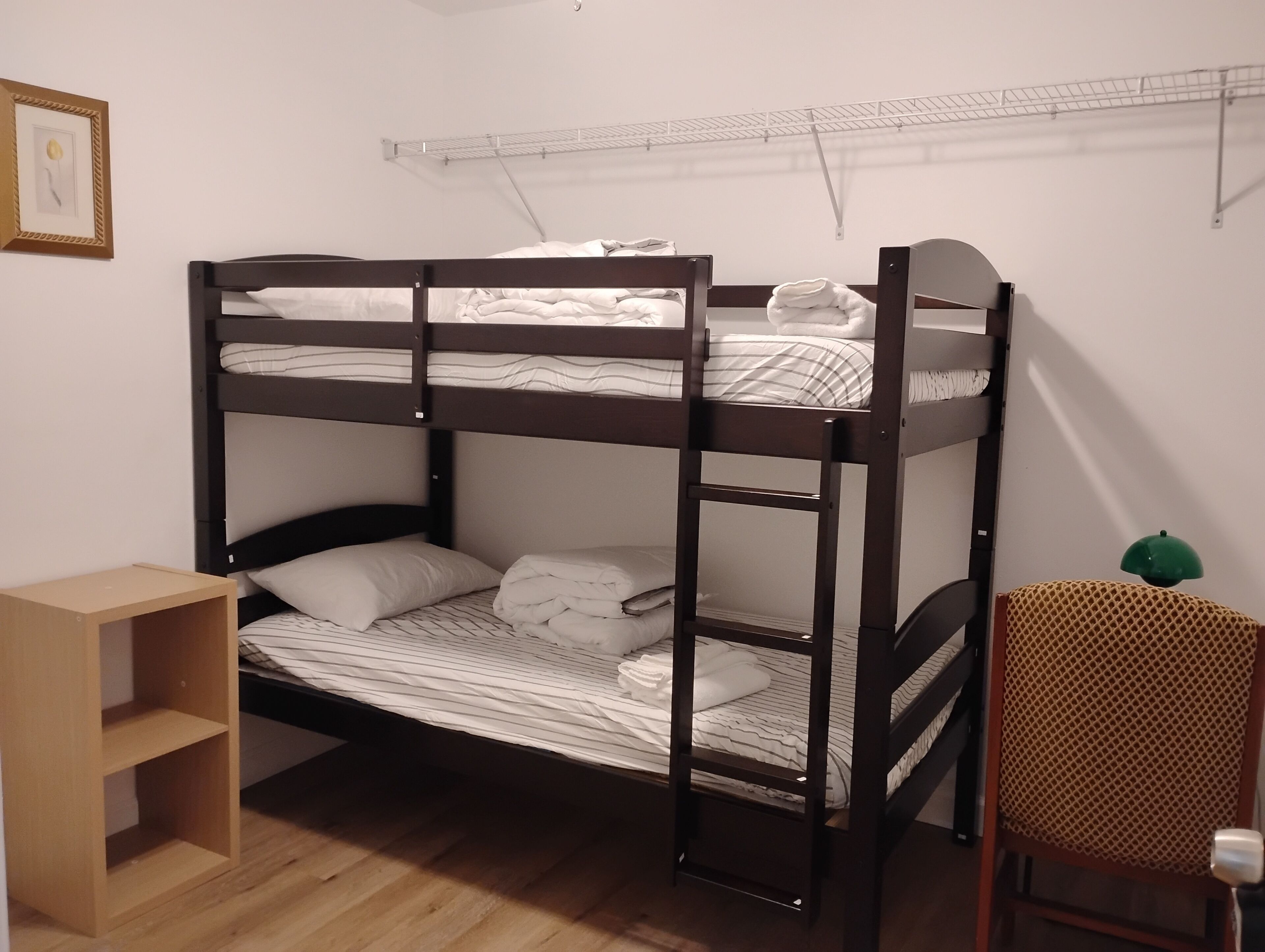 A bunk bed room with a wooden floor, a small wooden shelf, a green chair, and a white wall with a picture frame.