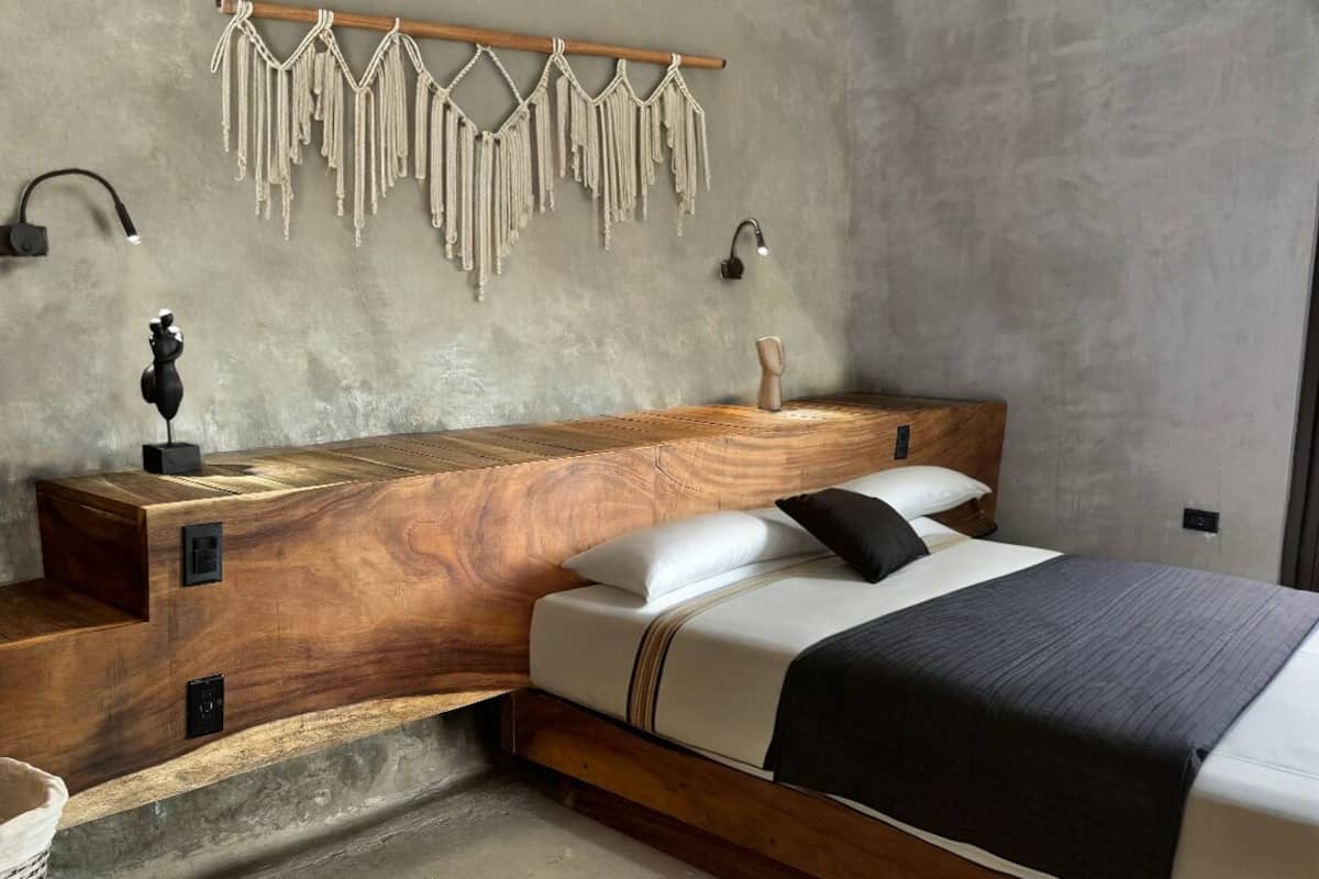 Une chambre moderne avec une tête de lit en bois, un lit avec une literie blanche et foncée, une table de chevet avec une lampe et des décorations murales en macramé.