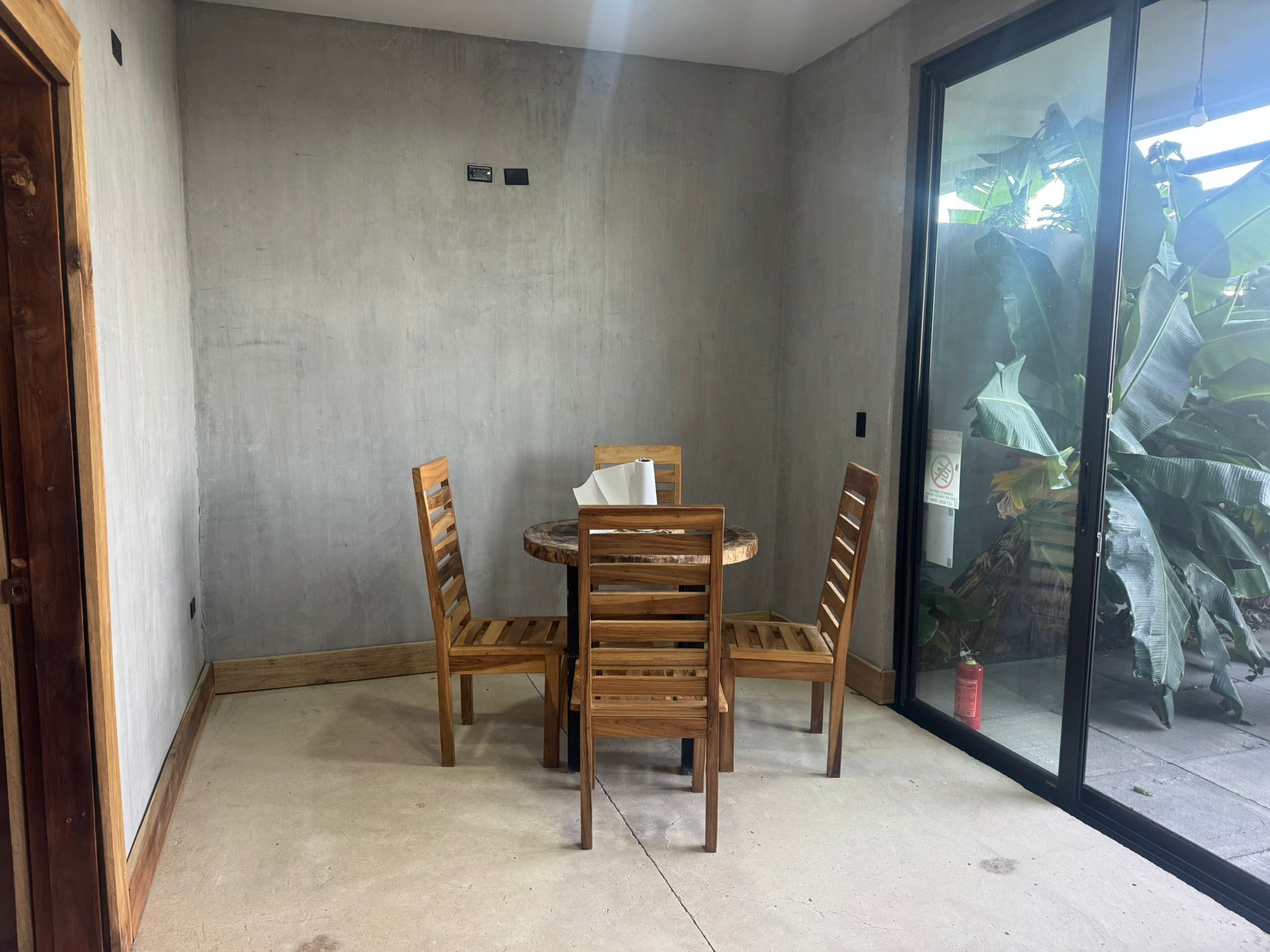 Un coin repas avec une table ronde et quatre chaises en bois, un mur en béton et une porte-fenêtre coulissante donnant sur un balcon agrémenté de plantes.