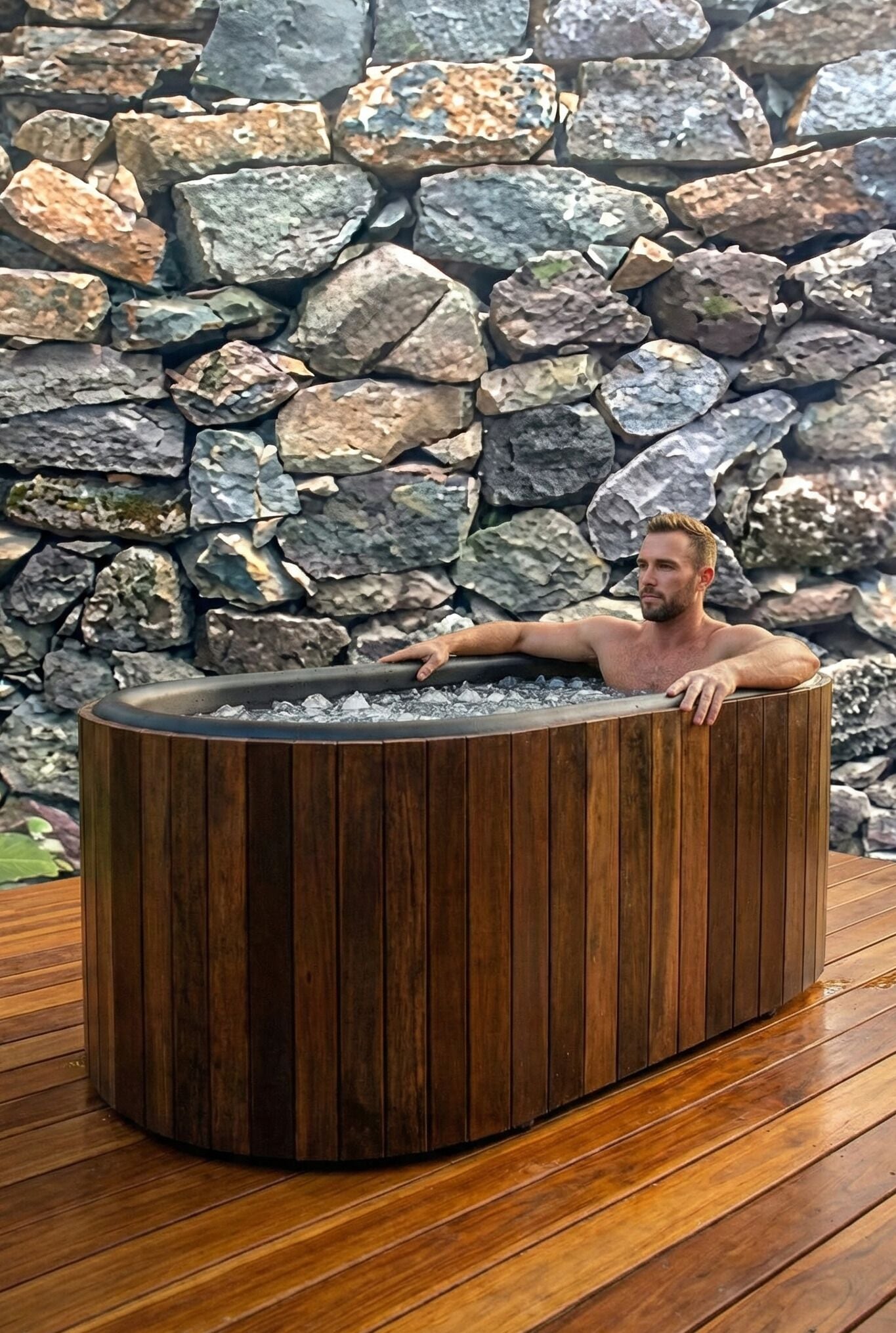 A man relaxing in a wooden hot tub with a stone wall backdrop.