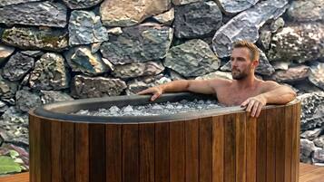 A man relaxing in a wooden hot tub with a stone wall backdrop.