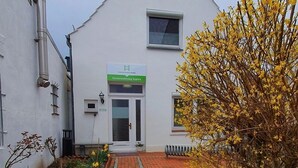 A building with a white facade, a brick pathway leading to the entrance, and a variety of flowers and plants around the entrance area.