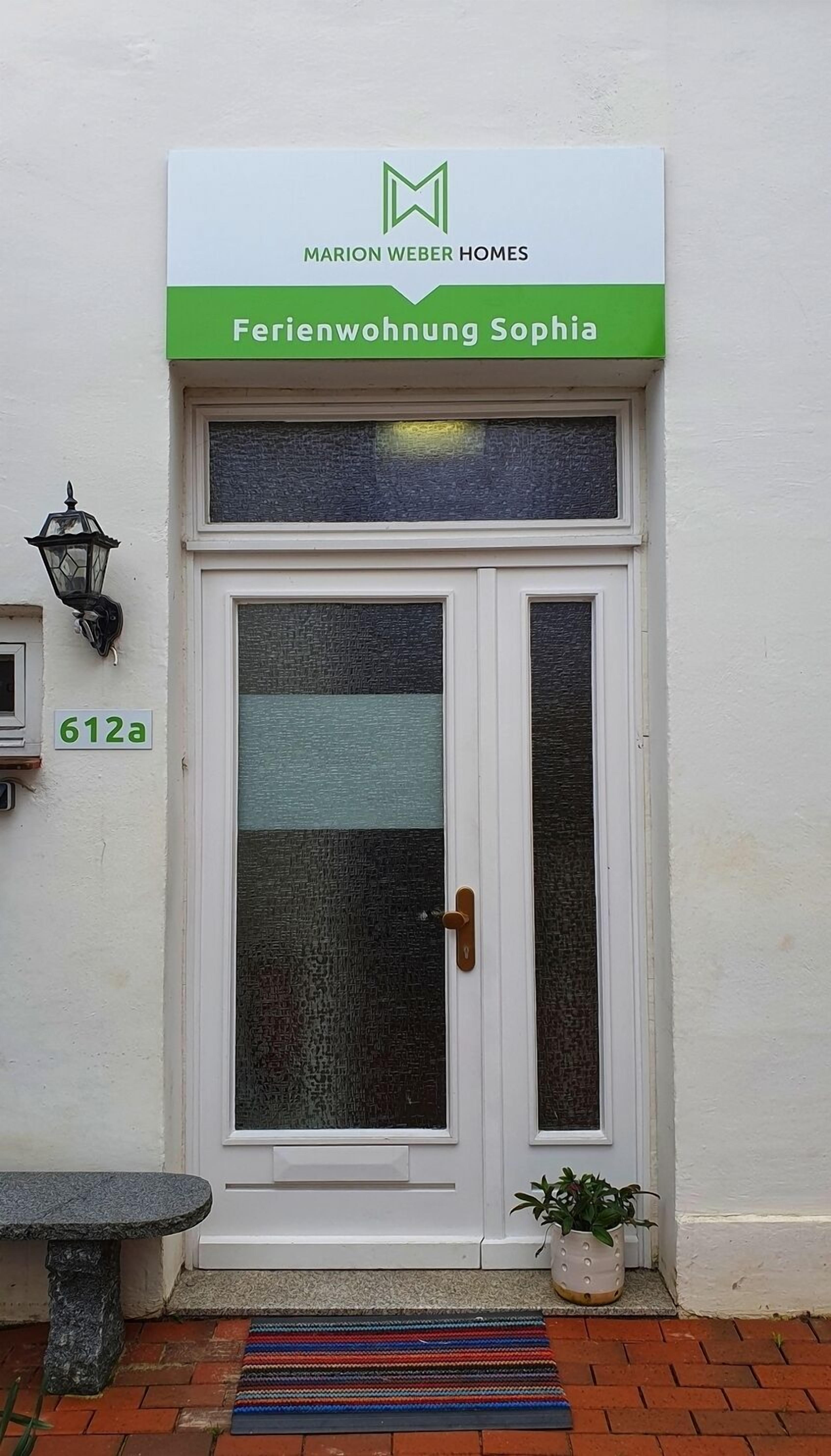 A white door with a green sign above it reading "Marion Weber Homes" and "Ferienwohnung Sophia". There is a number plate "612a" on the wall to the left of the door.