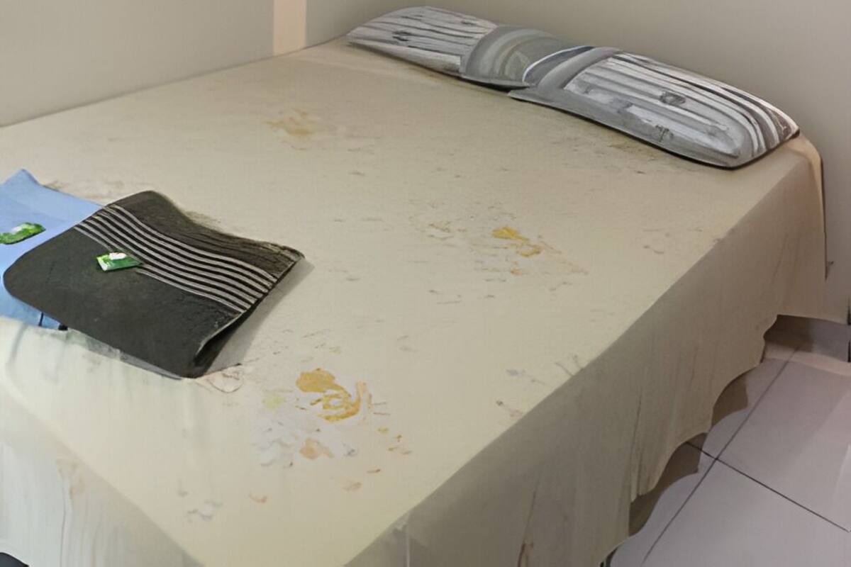 A small, single-bed room with a plain white wall, a light green mattress, and a pillow with a striped pattern. A black object, possibly a bag or a book, is on the bed.