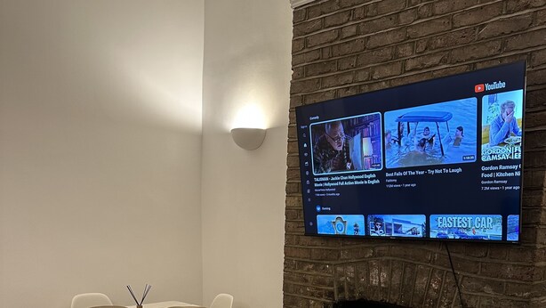A modern living room with a brick fireplace, a flat-screen TV mounted above it, and a dining area with a white table and chairs.