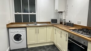 A compact kitchen with a washing machine, microwave, gas stove, and a window with blinds.
