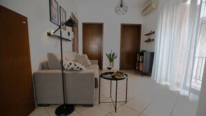 Sala de estar com sofá, luminária de chão, mesa pequena com um livro e planta em vaso. Há duas portas e uma janela com cortinas.