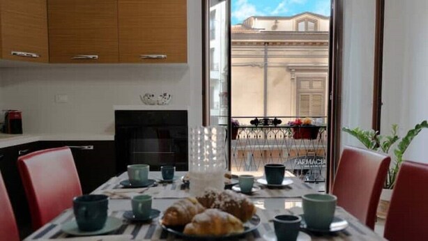 Uma área de refeições com uma mesa posta para o pequeno-almoço, incluindo croissants e chávenas, com vista para um edifício através de uma porta de vidro.