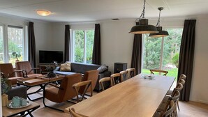 A spacious living room with a long wooden dining table, comfortable seating, a flat-screen TV, and large windows offering a view of greenery.