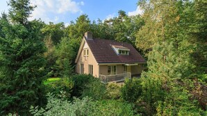 A small, single-story house with a sloped roof, surrounded by dense greenery and trees.